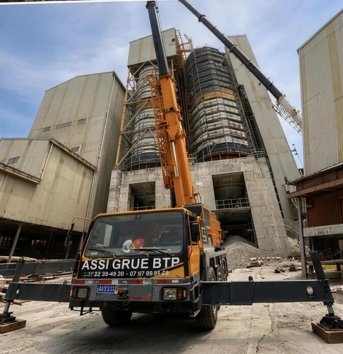 Grue ASSI GRUE BTP jaune calée devant une usine en hauteur